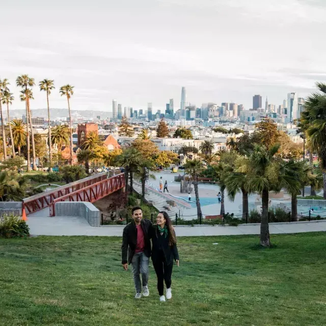 A couple walks toward the camera with Dolores Park and the San Francisco Skyline behind them.